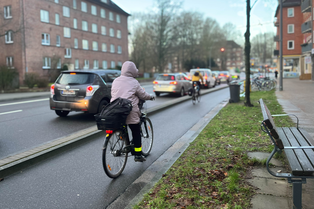 Radwege in Hamburg: Beispielbild für Protected Bikelanes. Eine Frau fährt auf einem Fahrradweg, der von der Hauptstraße mit einer Erhöhung ähnlich einem Bordstein abgetrennt ist. Auf der Straße fahren Autos, im Hintergrund rote Klinkerbauten.