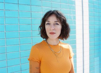 Antje Schomaker: „Leute, ich bin jetzt sexy” Antje Schomaker steht in einem orangenen Oberteil vor blauen Kacheln