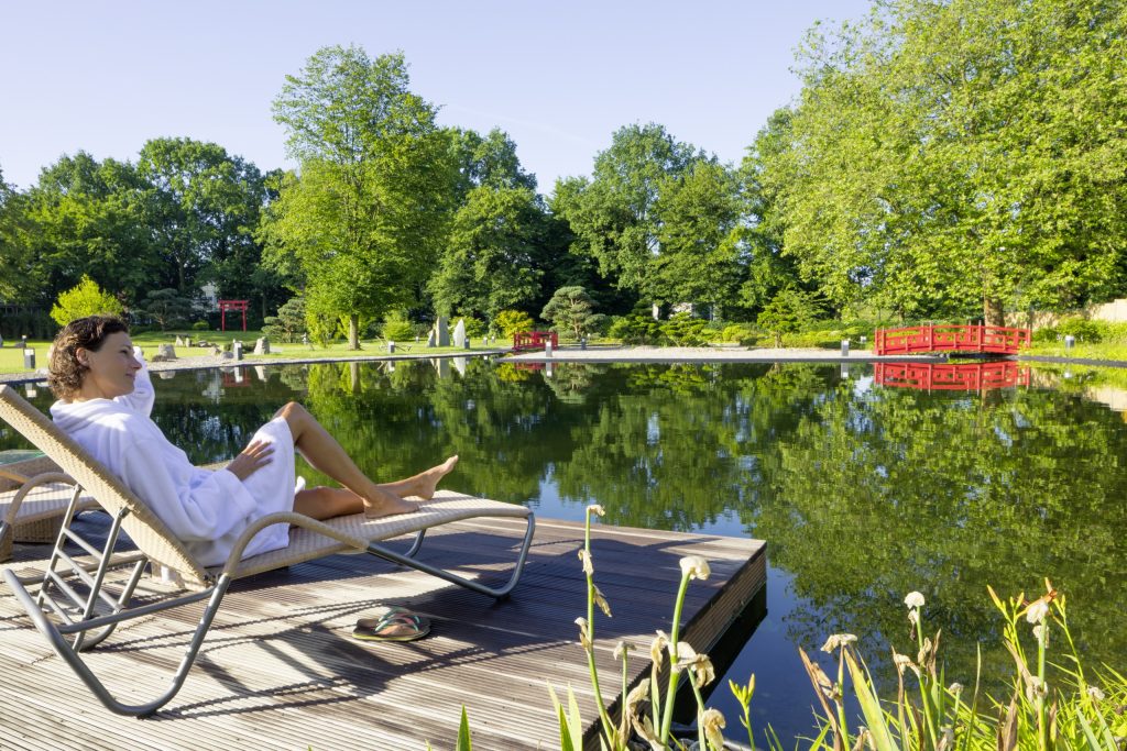 Freibäder Bäderland: Eine Frau sitzt auf einer Liege vor einem Teich. Im Hintergrund stehen Bäume.