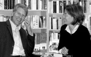 Buchhandlung Christiansen: Eine Schwarz-Weiß-Fotografie eines Paares. Links sitzt ein Mann, der lachend in die Kamera schaut. Rechts neben ihm sitzt eine Frau, die den Mann anlächelt. Im Hintergrund steht ein Bücherregal.