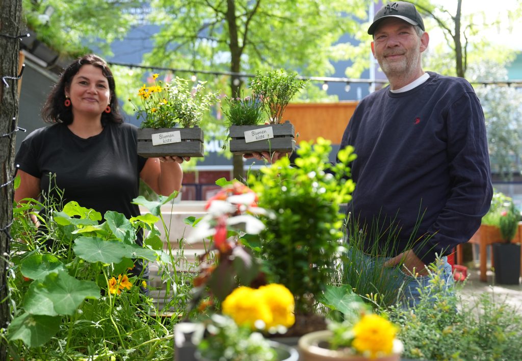 Eine Frau und ein Mann stehen vor zahlreichen Pflanzen. Sie halten jeweils einen kleinen Blumenkorb in der Hand.