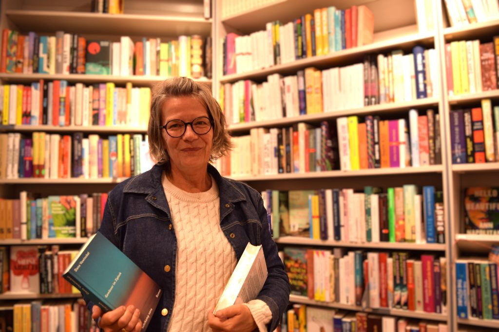 Buchhandlung Christiansen: Eine Frau mit weißem Pullover und einer Jeansjacke steht vor einem Bücherregal. Sie hält zwei Bücher in den Händen.