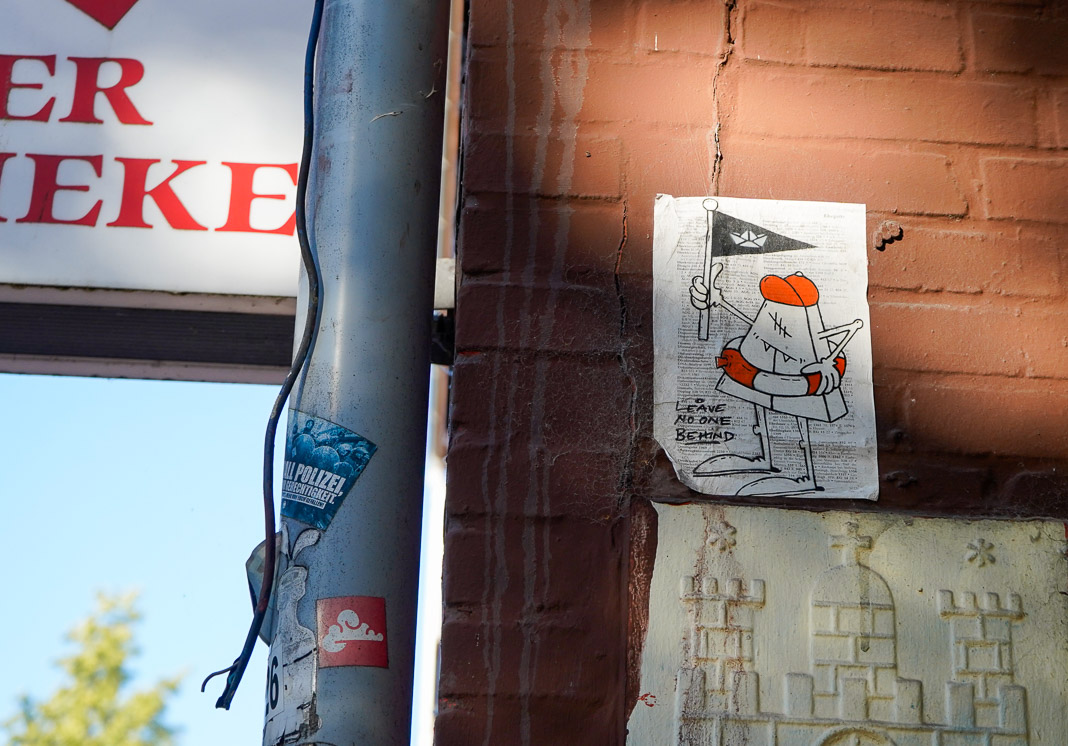 Motiv von HalloKarlo. Eine fiktive Figur mit roter Kappe. Es hält eine Flagge und trägt einen Rettungsring. Neben der Figur der Spruch "Leave No One Behind".