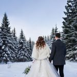 Hamburg heiratet gerne kalt Eine romantische Winterhochzeitsszene mit einem frisch verheirateten Ehepaar. Die Braut trägt ein elegantes weißes Winterhochzeitskleid mit langen Ärmeln, feinen Spitzendetails und einem mit Fell gefütterten Umhang. Der Bräutigam ist in einem klassischen dunklen Anzug mit einem warmen Wollmantel und einem weißen Ansteckblumenstrauß gekleidet. Sie stehen zusammen in einer schneebedeckten Landschaft, im Hintergrund verschneite Tannenbäume. Die Atmosphäre ist ruhig, mit sanftem Sonnenlicht, das durch den bewölkten Himmel bricht, und zarten Schneeflocken, die um sie herum fallen.