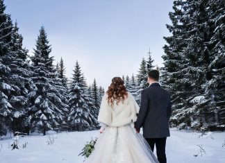Hamburg heiratet gerne kalt Eine romantische Winterhochzeitsszene mit einem frisch verheirateten Ehepaar. Die Braut trägt ein elegantes weißes Winterhochzeitskleid mit langen Ärmeln, feinen Spitzendetails und einem mit Fell gefütterten Umhang. Der Bräutigam ist in einem klassischen dunklen Anzug mit einem warmen Wollmantel und einem weißen Ansteckblumenstrauß gekleidet. Sie stehen zusammen in einer schneebedeckten Landschaft, im Hintergrund verschneite Tannenbäume. Die Atmosphäre ist ruhig, mit sanftem Sonnenlicht, das durch den bewölkten Himmel bricht, und zarten Schneeflocken, die um sie herum fallen.