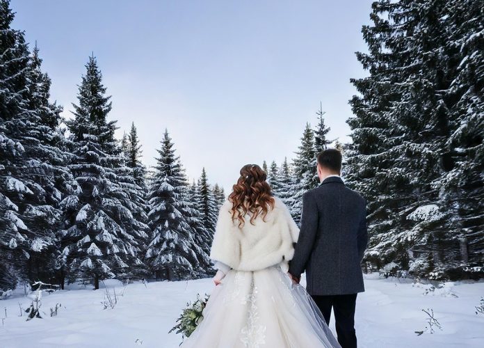 Eine romantische Winterhochzeitsszene mit einem frisch verheirateten Ehepaar. Die Braut trägt ein elegantes weißes Winterhochzeitskleid mit langen Ärmeln, feinen Spitzendetails und einem mit Fell gefütterten Umhang. Der Bräutigam ist in einem klassischen dunklen Anzug mit einem warmen Wollmantel und einem weißen Ansteckblumenstrauß gekleidet. Sie stehen zusammen in einer schneebedeckten Landschaft, im Hintergrund verschneite Tannenbäume. Die Atmosphäre ist ruhig, mit sanftem Sonnenlicht, das durch den bewölkten Himmel bricht, und zarten Schneeflocken, die um sie herum fallen.