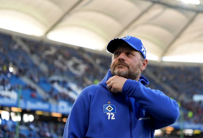 Der ehemalige HSV-Trainer Steffen Baumgart trägt eine Cap und eine Jacke mit dem HSV-Logo. Er steht in einem Fußballstadion.