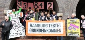 Hamburg testet Grundeinkommen übergibt die Stimmen an den Senat. Viele Menschen stehen zusammen und halten Schilder mit den Nummern 95.842 hoch. 