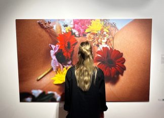 Lebefrauu: Feminismus nebenbei sexy machen Eine Frau steht vor einem Werk von Lebefrauu. Auf dem Werk sieht man eine Vulva mit Blumen. Die Person verdeckt den Intimbereich.