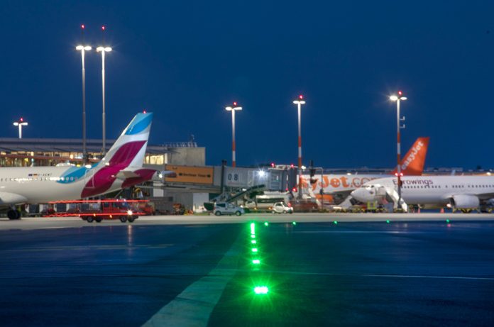 Ein Bild vom Flughafen Hamburg im Dunkeln. Man sieht verschiedene Flugzeuge, die auf dem Rollfeld stehen und bunte Lichter leuchten.