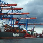 Landstrom wird am Hafen wenig genutzt Ein Bild vom Hamburger Hafen. Der Himmel ist grau-blau und stark bewölkt. Ein Containerschiff liegt vor rot-blauen Hafenkranen. Hier gibt es seit Mai Landstrom.