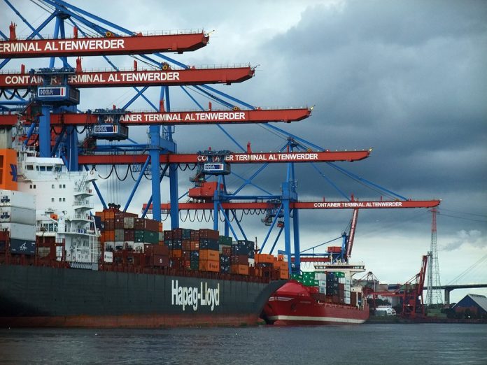 Ein Bild vom Hamburger Hafen. Der Himmel ist grau-blau und stark bewölkt. Ein Containerschiff liegt vor rot-blauen Hafenkranen. Hier gibt es seit Mai Landstrom.