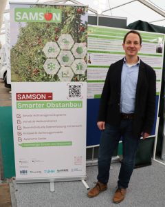 Auf der rechten Seite steht ein Mann in Jackett und Jeans. Links neben ihm steht ein großer Aufsteller, auf dem das Projekt Samson - Digitalisierung im Obstanbau erläutert wird. Foto: SAMSON-Projekt