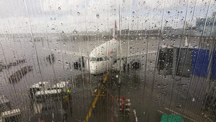 Hinter einer verregneten Scheibe steht ein Flugzeug an einem Flughafen.