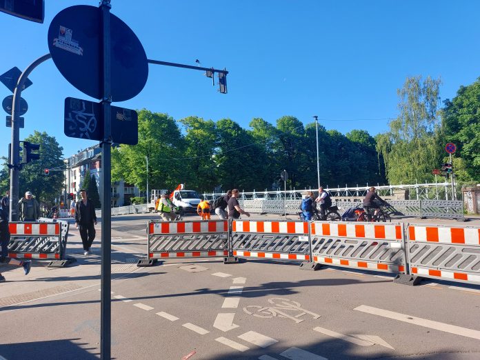 Ein Bild von der Wartenaubrücke bei blauem Himmel. Fußgänger laufen durchs Bild. Es ist eine Baustelle mit roten Absperrungen auf der Straße.