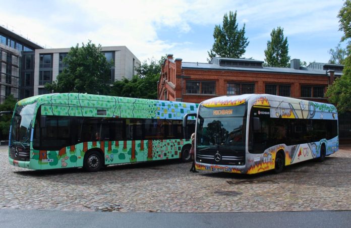 Die beiden HVV Paintbusse. Links ein grüner mit Pixel-Designs und rechts ein gelb-blauer Bus mit den Motiven Krieg und Frieden.
