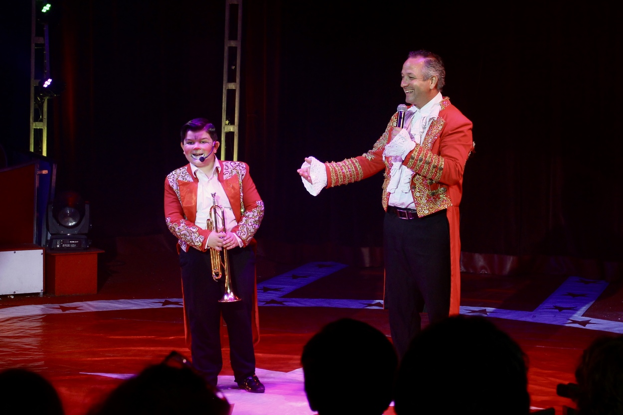 Ein Mann im Zirkusdirektor-Kostüm und ein Junge, der als Clown verkleidet ist, stehen in der Manege des Zirkus Frank. Der Junge hält eine Trompete in den Händen.
