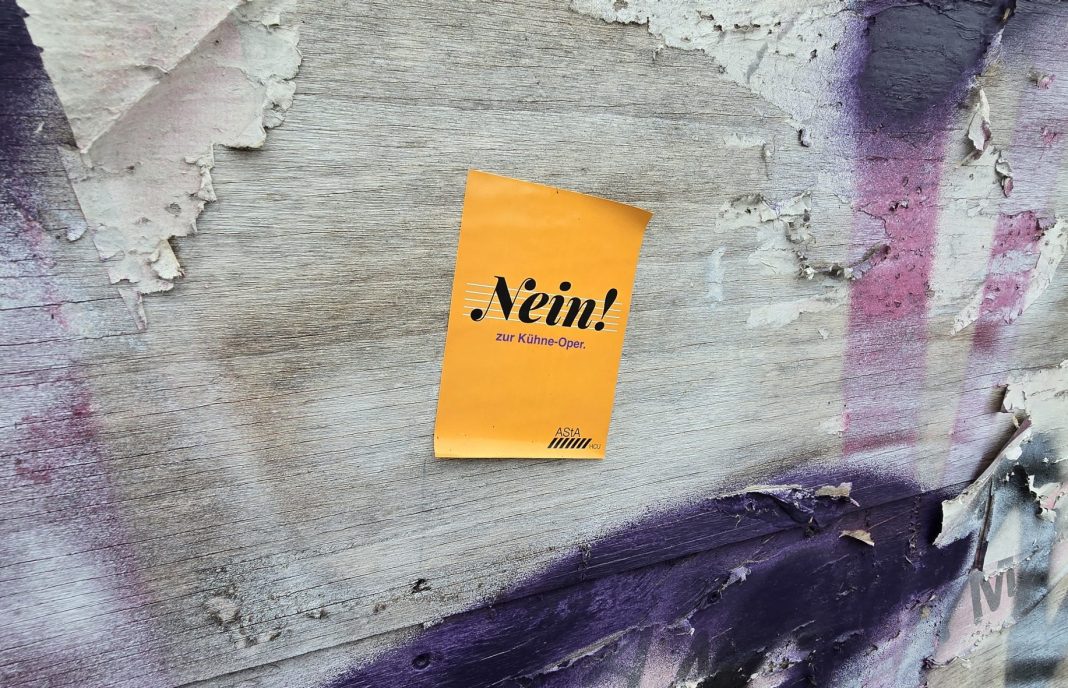 An einer mit Graffiti bemalten Wand ist ein gelber Sticker zu sehen. Auf diesem steht 