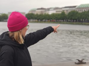 Konczalla zeigt mit ihrem Finger vom Jungfernstieg aus über die Binnenalster auf den Ballindamm und erkllärt wo beim Hamburg Ironman die Wechselzone aufgebaut wird. Sie trägt eine helle, pinke Mütze über ihren blonden Haaren, und eine schwarze Jacke.
