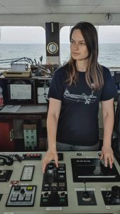 Lea Brakemeier steht auf der Brücke eines Küstenmotorschiffes. Sie blickt in die Ferne. Im Hintergrund sieht man das Meer.