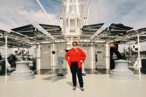Cynthia auf dem Deck der Cap San Diego. Sie trägt ein rotes Oberteil, steht mit den Armen in der Hüfte auf dem Schiff und guckt in die Kamera.