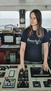 Lea Brakemeier steht auf der Brücke eines Küstenmotorschiffes. Sie blickt in die Ferne. Im Hintergrund sieht man das Meer.