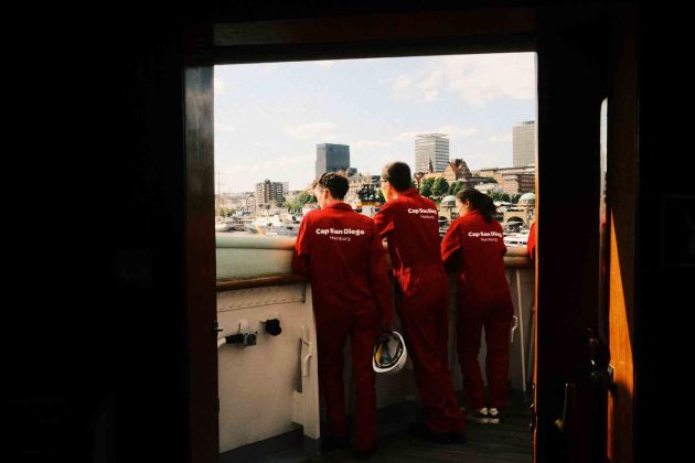 Drei junge ehrenamtliche Mitarbeitende der Cap San Diego stehen neben der Brücke an der Reling und schauen auf den Hamburger Hafen.