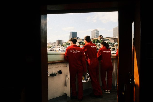 Drei junge ehrenamtliche Mitarbeitende der Cap San Diego stehen neben der Brücke an der Reling und schauen auf den Hamburger Hafen.