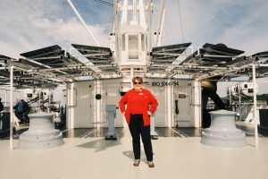Cynthia auf dem Deck der Cap San Diego. Sie trägt ein rotes Oberteil, steht mit den Armen in der Hüfte auf dem Schiff und guckt in die Kamera.