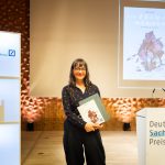 Ulli Lust gewinnt Deutschen Sachbuchpreis 2025 Die Gewinnerin des Deutschen Sachbuchpreises steht mit Buch und Urkunde in der Hand auf einer Bühne