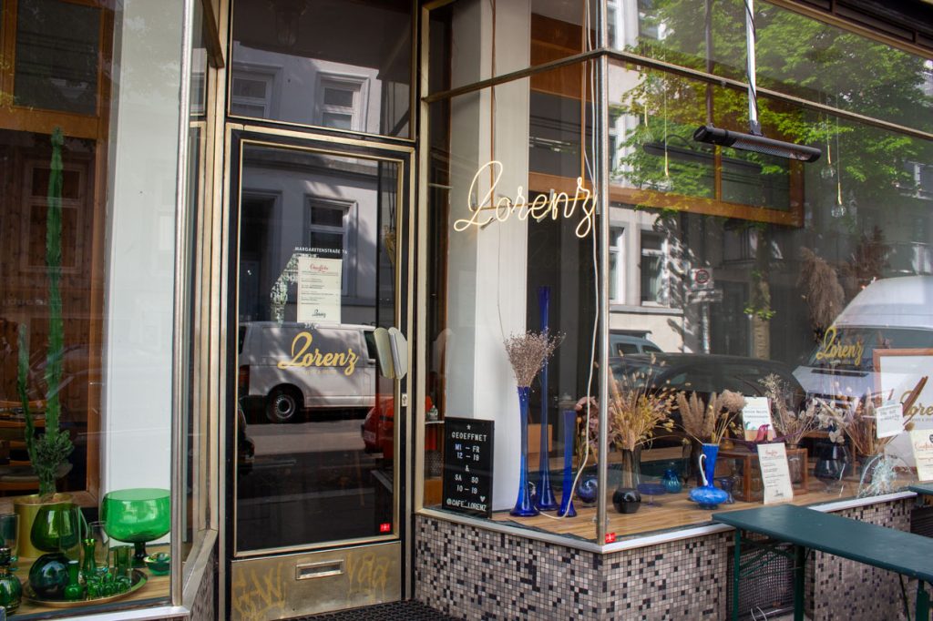 Das Café Lorenz von Außen. Eine helle Leuchtschrift formt den Namen Lorenz rechts neben der Eingangstür. Rechts und links sind Große Fenster. Auf den Fensterbänken stehen Grüne und blaue Vasen mit Trockenblumen.