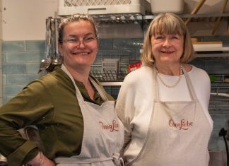 Oma Liebe Lunch Club: Eine kulinarische Reise in die Vergangenheit Zwei Frauen stehen nebeneinander in einer Küche. Sie tragen eine beige Schürze und lächeln.