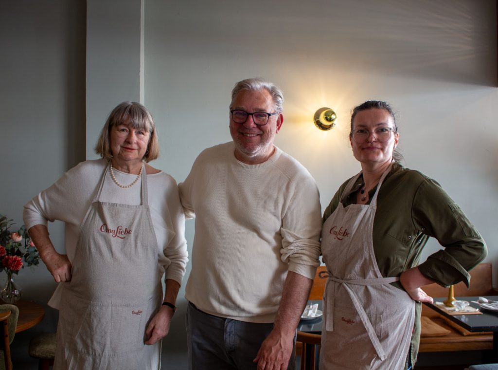 Drei Personen stehen nebeneinander vor einer hellgrauen Wand im Café Lorenz. In der Mitte steht ein Mann mit einem grauen Pullover, rechts und links von ihm steht jeweils eine Frau. Diese tragen eine beigen Schürze mit der Aufschrift Oma Liebe.