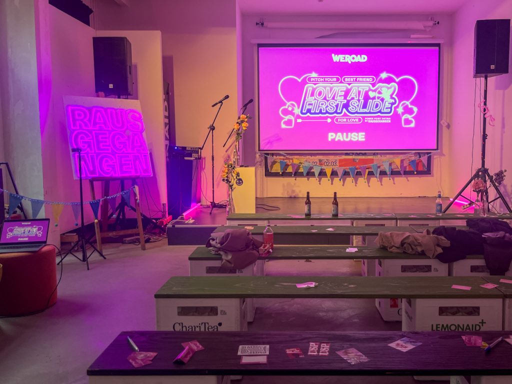 Ein pinker Bildschirm auf der Bühne mit der Aufschrift "Love at First Slide". Leere Bänke und ein Schild rechts mit der Aufschrift "Rausgegangen".