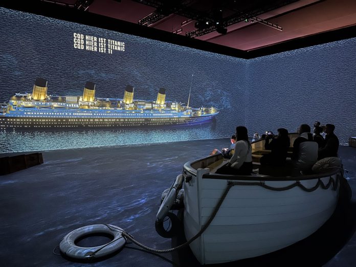 360-Grad-Lichtprojektion in einem großen Raum. Sie zeigt die Titanic. In der Mitte steht ein Rettungsschiff, in dem sich Besucher*innen befinden.