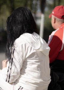 Eine Frau mit langen, schwarzen Haaren sitzt in einem weißen Trainingsanzug neben einem Mann in einer roten Baseballcap und einer roten Trainingsjacke. Beide Personen sieht man nur von hinten.