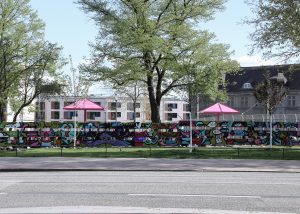 Es ist ein Sichtschutz zu sehen, der mit buntem Graffiti besprüht ist. Davor ist eine grüne Wiese und dann eine Straße.