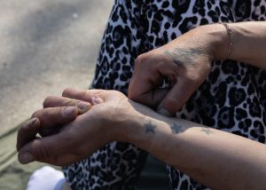 Nahaufnahme eines Handgelenks, auf dem drei Sterne tättowiert sind. Auf der anderen Hand ist ein weiteres Tattoo zu sehen. Die Person trägt eine Hose mit Leo Print.