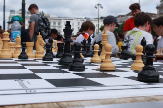 Schachfiguren im Vordergrund auf einem Schachbrett. Im Hintergrund sitzen Kinder und spielen in Reihen Schach