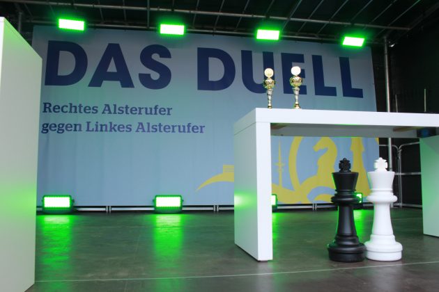 Die Bühne des Turniers ist mit grünem Licht beleuchtet. Auf einem Tisch stehen zwei kleine Pokale. Davor zwei überdimensionierte Schachfiguren.