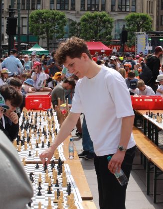 Schach-Großmeister Luis Engel spielt an vielen Schachbrettern gegen Schülerinnen und Schüler. Er trägt ein weißes Shirt und hat eine Wasserflasche in der Hand.