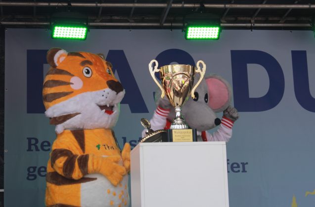 Auf einer Bühne mit grünem Licht stehen zwei Menschen in Maskottchenkostümen eines Tigers und einer Maus. Davor steht auf einem Podest ein großer goldener Pokal.