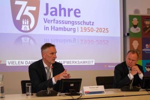 Zwei Männer sitzen an einem Tisch. Sie tragen schwarze Anzüge und stellen den Verfassungsschutzbericht vor.