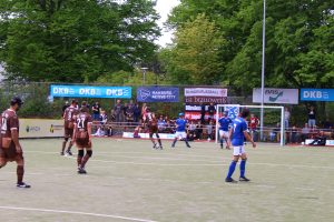 Blindenfußballer bei einem Spiel, im Hintergrund Werbebanner und Bäume. 