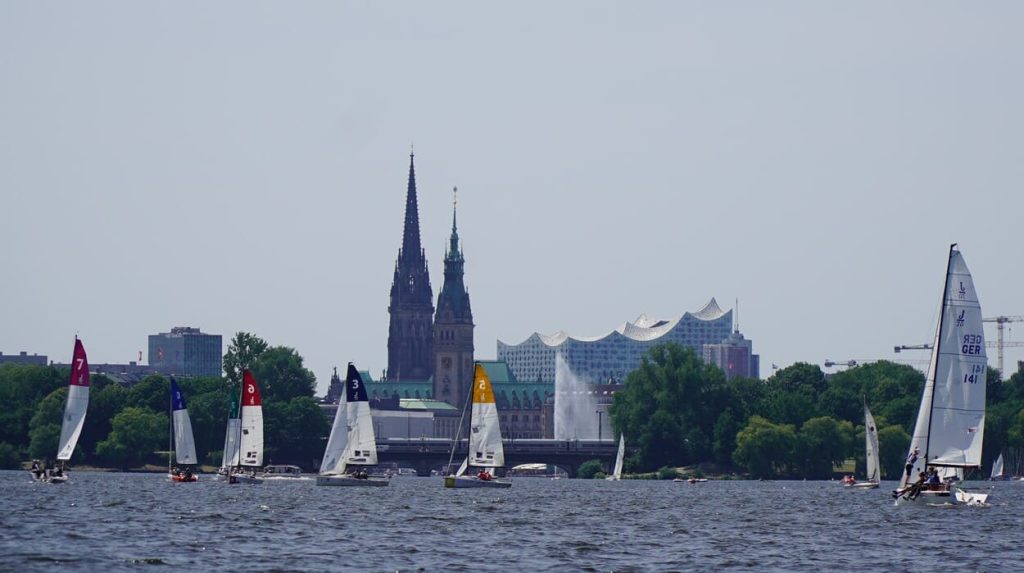 Segelboote beim Helga Cup auf der Außenalster. Im Hintergrund sieht man das wellige Dach der Elbphilharmonie, das Rathaus und den Turm der Nikolaikirche.