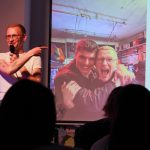 „Love at First Slide”: Pitch your Best Friend Ein junger Mann steht auf einer Bühne vor Publikum. Er deutet auf eine Präsentation neben ihm, die ein Foto von ihm und seinem besten Freund Arm in Arm zeigt.