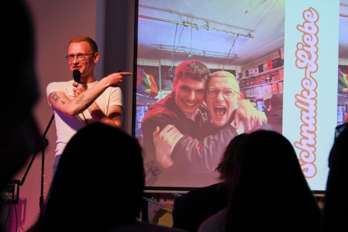 Love at First Slide Ein junger Mann steht auf einer Bühne vor Publikum. Er deutet auf eine Präsentation neben ihm, die ein Foto von ihm und seinem besten Freund Arm in Arm zeigt.