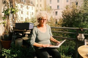 Magdalene Mintrop sitzt in der Sonne und blättert auf einem Balkon durch ein Fotobuch mit Bildern aus ihrer Zeit am Hamburger Hafen. Sie ist umgeben von Pflanzen, trägt eine Brille und ein olivfarbenes Oberteil. FINK.HAMBURG spricht mit ihr über: Frauen im Hamburger Hafen