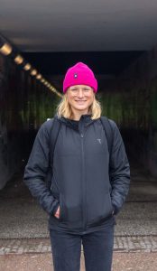 Konczalla steht lächelnd vor dem Eingang eines Tunnels, durch den beim Hamburg Ironman die Laufstrecke verläuft. Sie trägt eine helle, pinke Mütze über ihren blonden Haaren sowie eine schwarze Hose und Jacke und hat ihre Hände in den Jackentaschen.