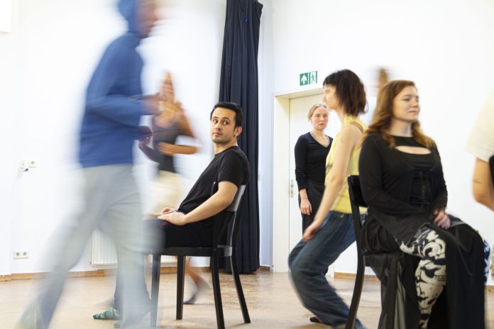 Lernende an der Schule für Schauspiel Hamburg gehen in einem Kursraum umher. Zwei sitzen in der Mitte des Raumes auf einem Stuhl
